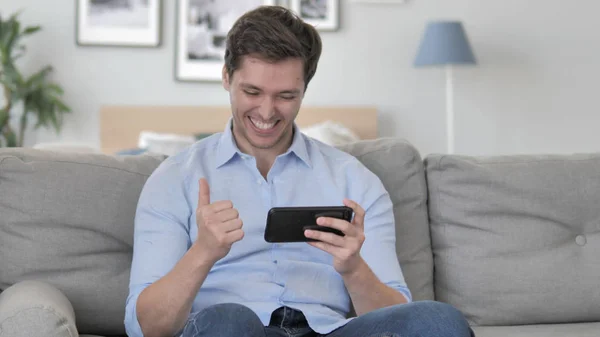 Hezký mladý muž jásající úspěch na smartphone při sezení na gauči — Stock fotografie