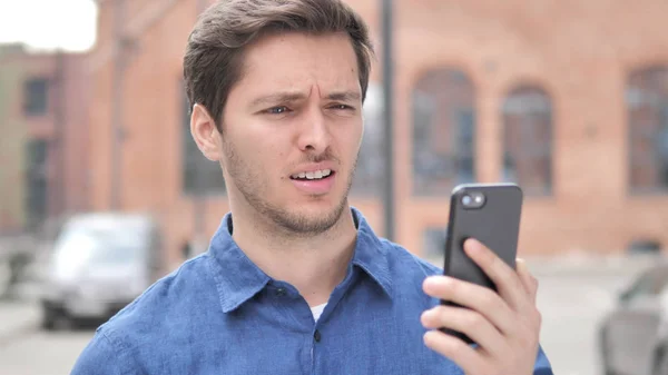 Retrato al aire libre de hombre joven molesto por la pérdida mientras se utiliza el teléfono inteligente — Foto de Stock