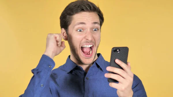 Hombre joven casual celebrando el éxito mientras usa el teléfono inteligente en el fondo amarillo — Foto de Stock