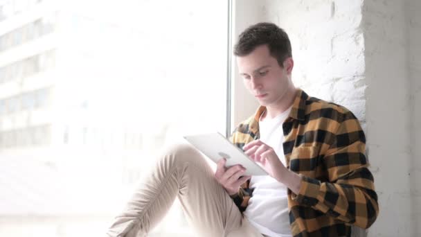 Junger Mann benutzt Tablet während er im Fenster sitzt — Stockvideo