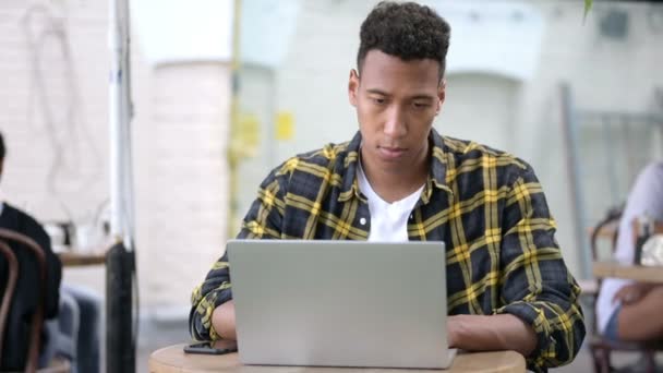 Молодой африканский человек с головной болью с помощью ноутбука, открытое кафе — стоковое видео