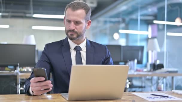 Молодой бизнесмен с помощью смартфона и ноутбука в офисе — стоковое видео