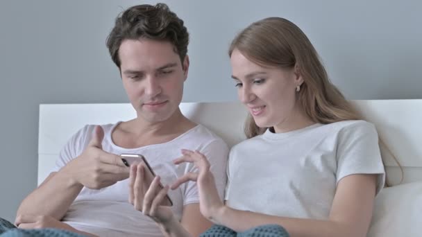 Fröhliches Paar sitzt im Bett und nutzt Smartphone — Stockvideo