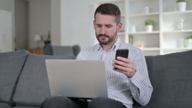 Bilgisayarlı genç adam evde akıllı telefondan konuşuyor. 