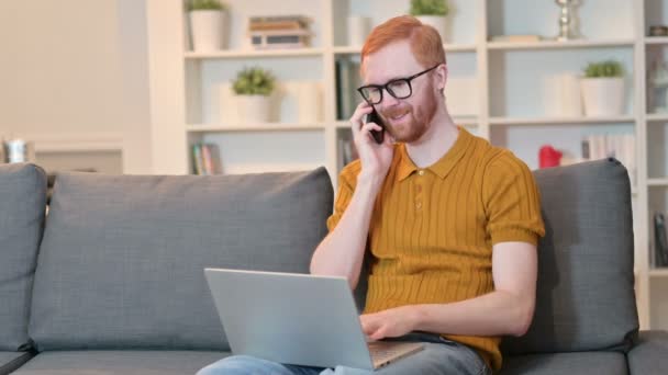 Vöröshajú férfi laptoppal beszél okostelefonon otthon — Stock videók