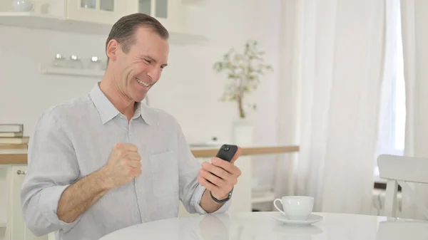 Middle Aged Man Celebrating Success on Smartphone at Home — Stock Photo, Image