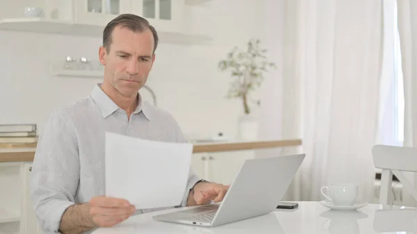 Hombre de mediana edad enfocado que trabaja en el ordenador portátil y el documento en casa — Foto de Stock