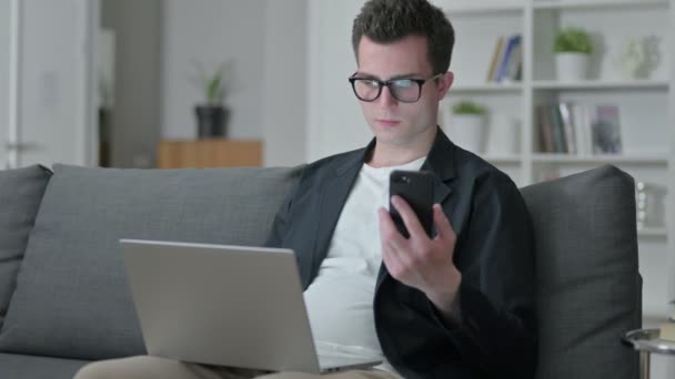 Diseñador masculino joven usando Smartphone y Laptop en casa — Vídeo de stock