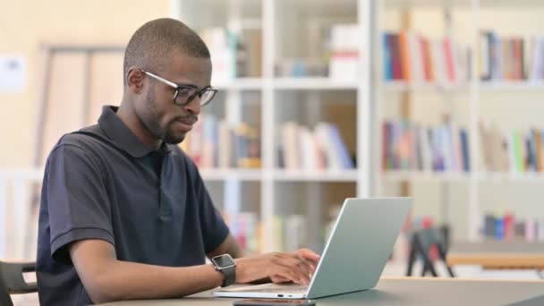 Jonge Afrikaanse man met laptop op zoek naar camera in de bibliotheek — Stockvideo