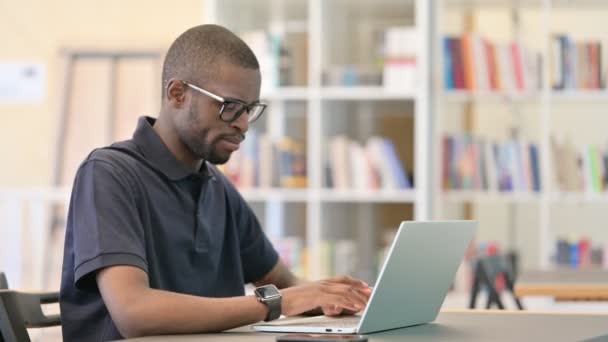 Junger Afrikaner mit Laptop lächelt in Bibliothek in die Kamera — Stockvideo
