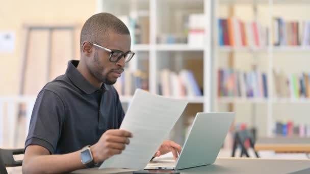 Jonge Afrikaanse man met laptop doet papierwerk in de bibliotheek — Stockvideo