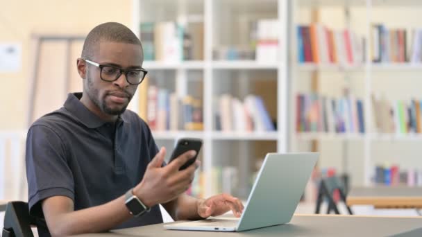 Gericht Afrikaanse Man werkt aan Smartphone en Laptop in de bibliotheek — Stockvideo