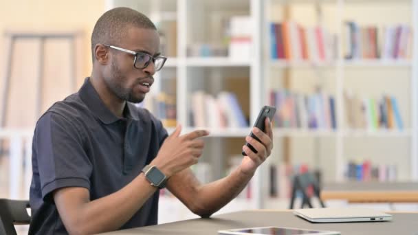 Perturbado hombre africano frente a la pérdida en el teléfono inteligente en la biblioteca — Vídeo de stock