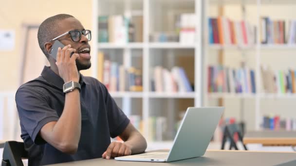 Junger Afrikaner mit Laptop spricht in Bibliothek mit Smartphone — Stockvideo
