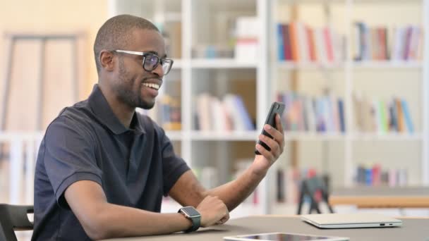 Vrolijke Afrikaanse man doet Video Call op Smartphone in de bibliotheek — Stockvideo