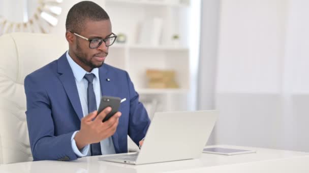 Empresario africano enfocado usando el ordenador portátil y el teléfono inteligente en la oficina — Vídeo de stock