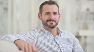 Genç adam koltukta oturuyor ve kameraya gülümsüyor. 