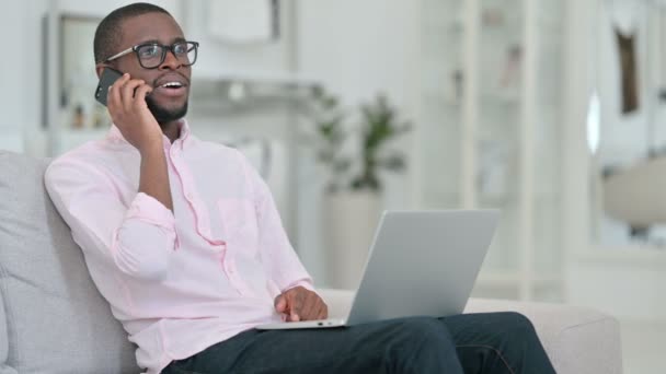Afrikansk man med bärbar dator Pratar på smartphone hemma — Stockvideo