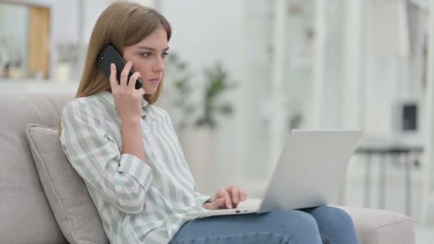 Mujer joven con el ordenador portátil hablando en el teléfono inteligente en casa — Vídeo de stock