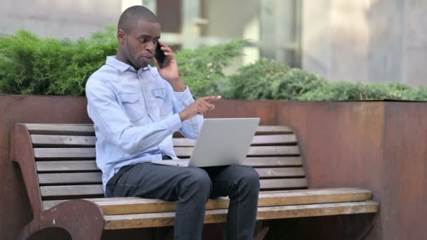 Afrikaanse man met laptop Praten op Smartphone Outdoor — Stockvideo