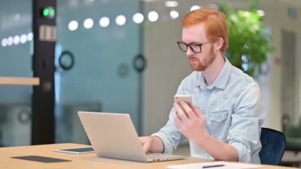 Ofiste akıllı telefon ve dizüstü bilgisayar kullanan sıradan bir kızıl adam. — Stok video