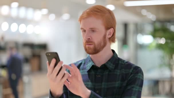スマートフォンで成功を祝うひげの赤毛の男の肖像 — ストック動画