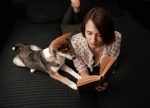 Tyttö Koiran Kanssa Makaa Mustassa Sängyssä Lukee Kirjaa — kuvapankkivalokuva