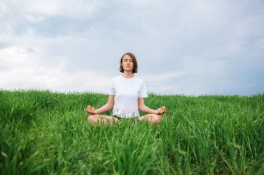 Genç bir kız çimlerin üzerinde otururken meditasyon yapıyor. Doğadaki kız. Ön görünüm