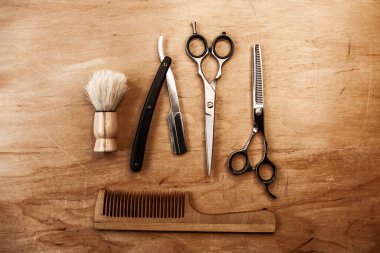 İki makas, tehlikeli bir jilet, bir fırça ve ahşap bir tarağın ortasında, ahşap bir masanın üzerinde yatıyor. Ahşap arka planda berber aracı.