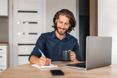 Bir adam bir kupa tutar ve bir deftere yazar. Evde çalışan bir adam. Gülümseyen adam