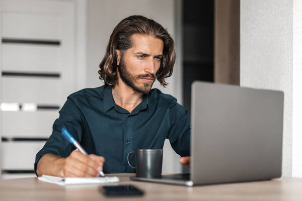 Portrait of a focused young man with long hair working remotely at a computer. A man writes information in a notebook and looks into a laptop