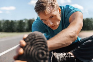 Atletik genç adam koşu bandında oturup sıkı bir antrenmana hazırlanırken bacaklarını esnetir. Yakın plan portre