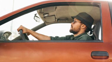 Sakallı ve güneş gözlüklü genç bir adam bir arabanın direksiyonunda oturur ve bir yolculuğa çıkar.