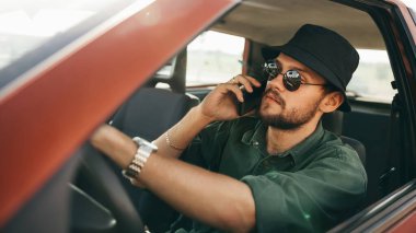 Güneş gözlüklü genç adam arabada otururken cep telefonuyla konuşuyor.