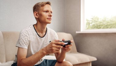 Genç sarışın adam oyun kumandasını tutarken konsolda oynuyor.