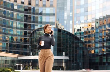 Genç gülümseyen bir iş kadını, elinde kahveyle şehrin iş yerinde yürüyor ve bir iş merkezinin arka planında akıllı bir telefonla konuşuyor.