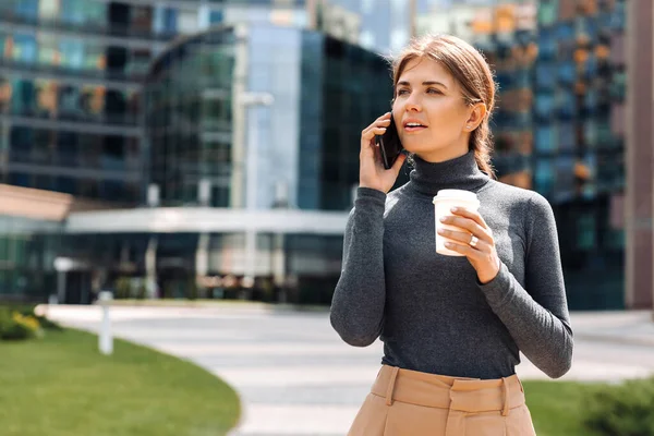 Genç bir iş kadını cep telefonuyla konuşuyor, elinde kahve tutuyor, şehrin iş yerinde duruyor ve bir iş merkezinin arka planına yaslanıyor.