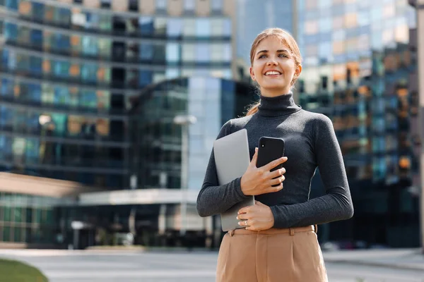 Portrait of a young blonde business woman with a smile holding a laptop and a mobile phone in her hands, stands on the street, in the business part of the city, against the background of a business center