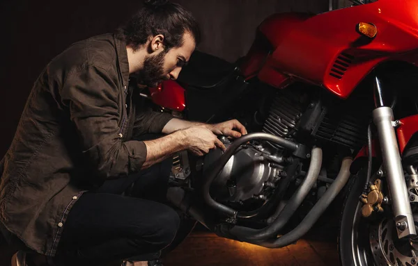 Sakallı bir adam garajda motosikletini tamir ediyor.