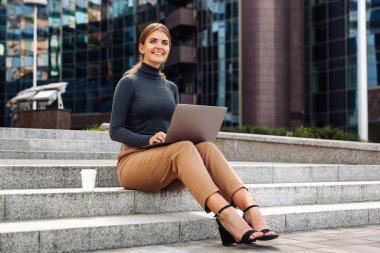 İş kadını iş merkezinin önündeki merdivenlerde otururken dizüstü bilgisayarda çalışıyor.