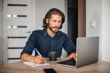 Bir adam dizüstü bilgisayarda çalışır ve bir deftere yazar. Adam internetten bir web sitesini izliyor.