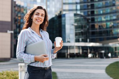 Yetişkin bir iş kadını elinde bir fincan kahve ve dizüstü bilgisayarla bir iş merkezinin arka planında durur ve kameraya gülümseyerek bakar.