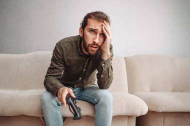 Üzgün bir adam elinde joystick ile kanepede oturuyor. Adam bir video oyunu kaybettiği için üzgün.