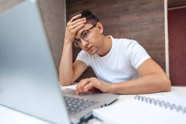 Evde dizüstü bilgisayarda çalışan gözlüklü yorgun genç adam.