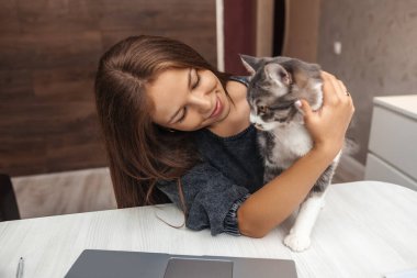 Genç kız masada otururken kedisine sarılıyor.