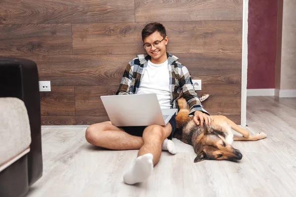 Evde dizüstü bilgisayarda çalışan ve yerde oturup köpeği okşayan mutlu genç adam.