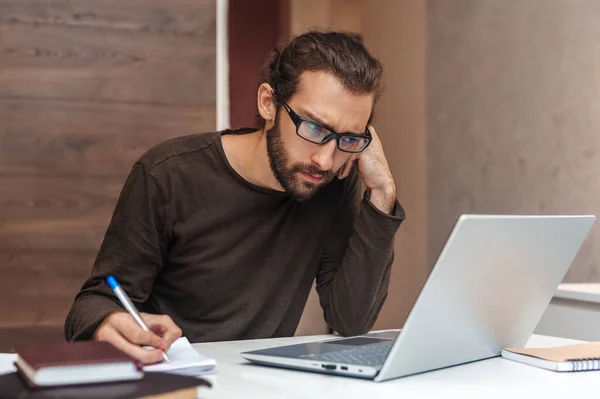 Joven Serio Con Barba Mira Portátil Escribe Bloc Notas — Foto de Stock