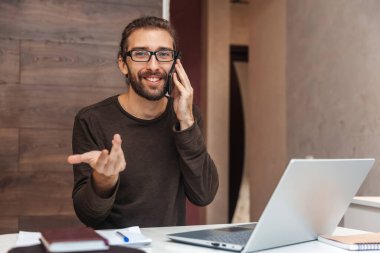 Evdeki çalışma masasında oturan mutlu adam cep telefonuyla konuşuyor.