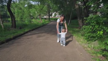 Mutlu, genç anne şirin bebeğe yürümeyi öğretiyor. Sevimli çocuk ilk adımlarını atıyor. Yeşil bahçede oğluyla birlikte yürüyen bir kadın. Aile kavramı, çocukluk, mutluluk, ebeveynlik, yaz