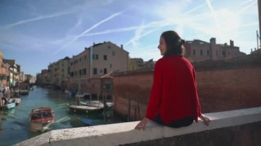 Köprünün kenarında oturan kırmızı kazaklı, çekici esmer kızın arkadan görünüşü süzülen gondollara ve kanal kenarında yürüyen turistlere bakıyor. İtalya 'da güneşin sıcacık ışınlarında dinlenmek.